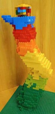 Side view of a twisting structure made of legos that transitions from dark green at the bottom to light green, yellow, orange, red, and then blue on top. The blue legos are shaped like a hand that's cupping a yellow cube.