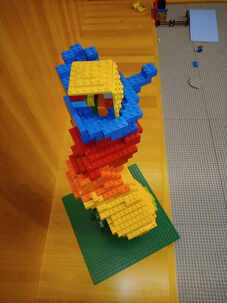 Top view of a twisting structure made of legos that transitions from dark green at the bottom to light green, yellow, orange, red, and then blue on top. The blue legos are shaped like a hand that's cupping a yellow cube.